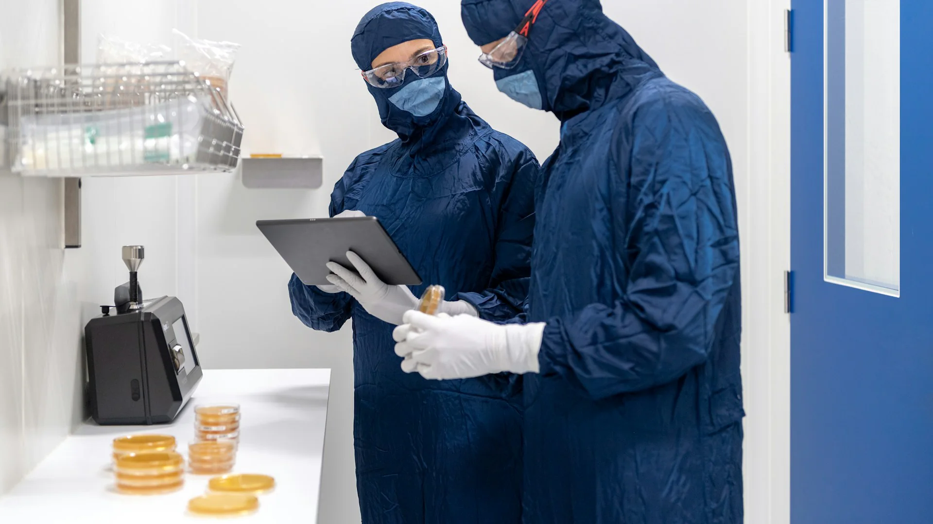 Two technicians in cleanroom gowns, masks and goggles reviewing a tablet beside a bench of culture plates